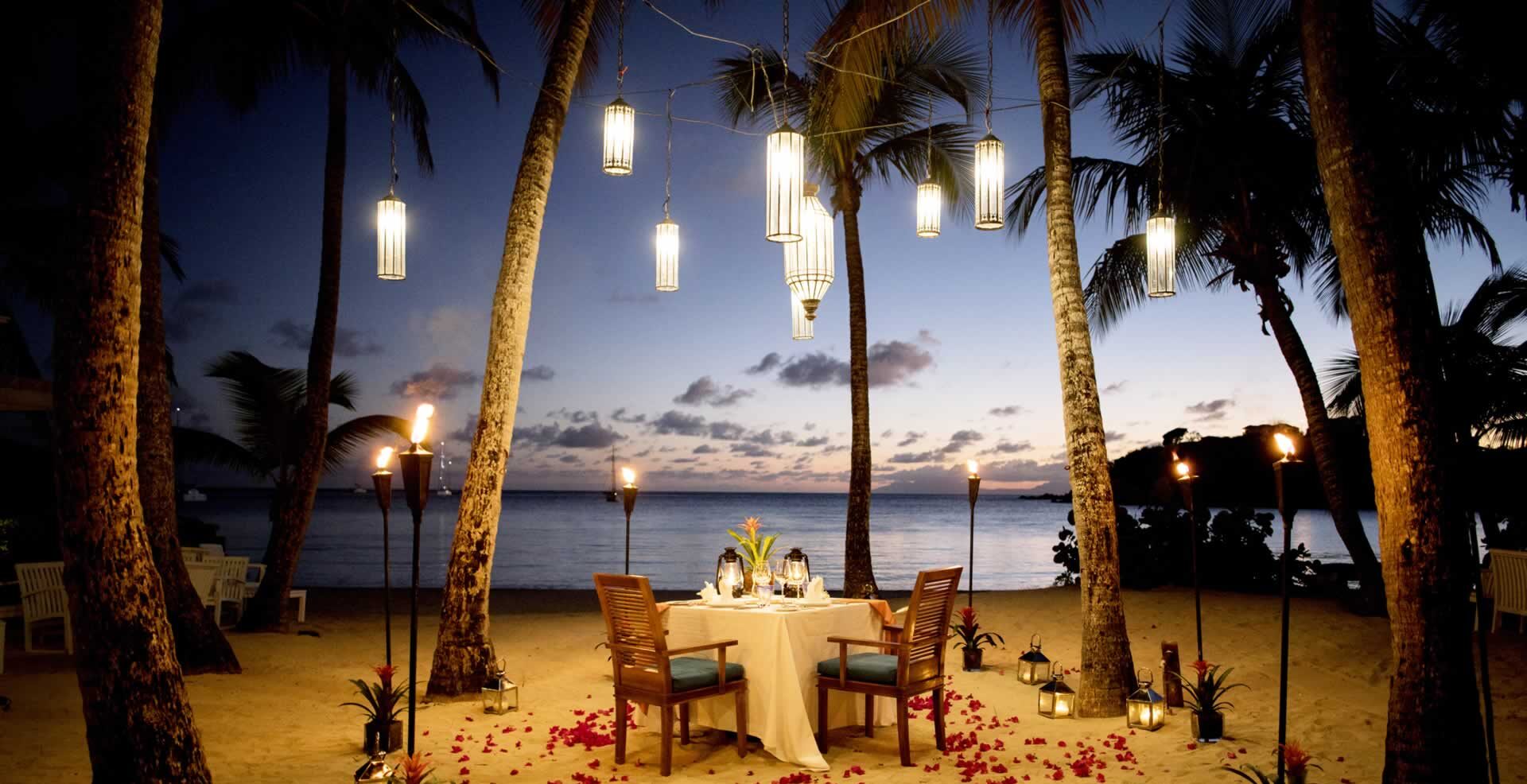 An intimate dinner setting with candle lit lamps and petals. On the beach in the evening.